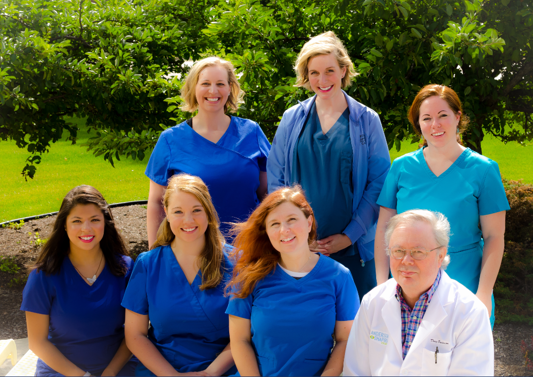 Seven techs in medical garb stand or sit in front of a green tree for a group photo